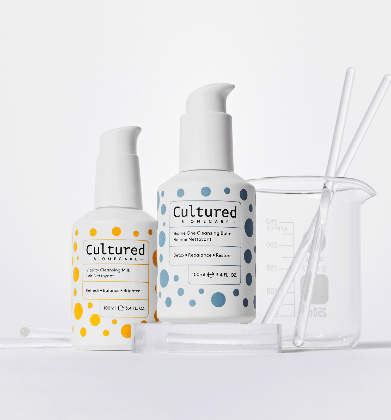 Vitality Cleansing Milk and Biome One Cleansing Balm skincare bottles in front of clear lab glassware & white background.